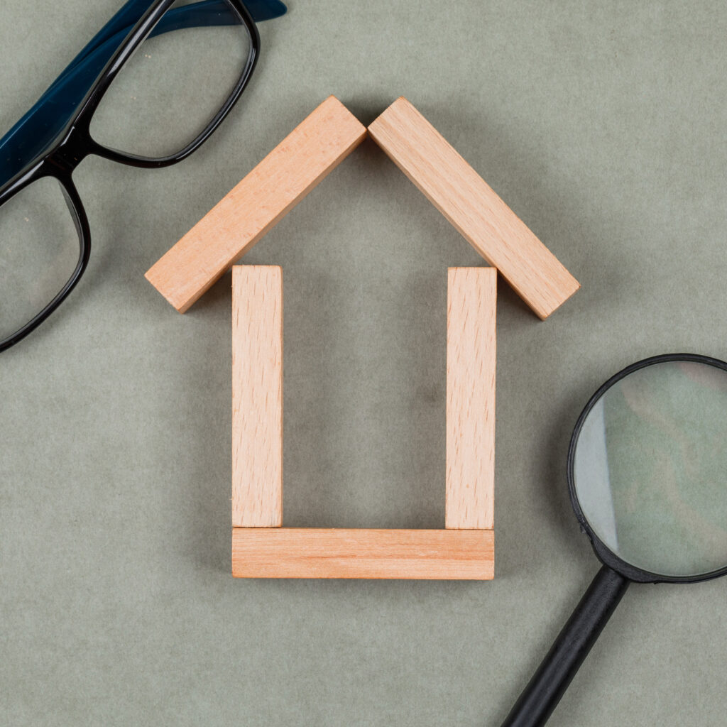 Real estate concept with house made of wooden blocks, glasses, magnifying glass on grey background close up.